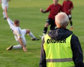 Ein Sicherheitsmitarbeiter in einer gelben Weste beobachtet ein Fußballspiel, während ein Spieler auf dem Rasen gefallen ist. Im Hintergrund sind weitere Spieler in roten und weißen Trikots zu sehen.