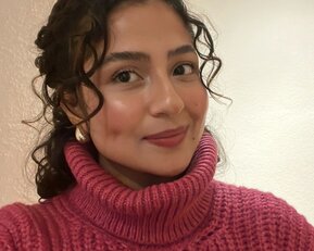 A person with curly hair is wearing a pink turtleneck sweater and smiling at the camera. The background is neutral.