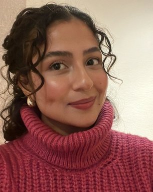 Eine Person mit lockigem Haar trägt einen rosa Rollkragenpullover und lächelt in die Kamera. Der Hintergrund ist neutral.