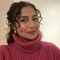 A person with curly hair is wearing a pink turtleneck sweater and smiling at the camera. The background is neutral.