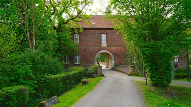Ein historisches Gebäude mit einem großen Torbogen, umgeben von Bäumen und Sträuchern, an einer gepflasterten Straße.