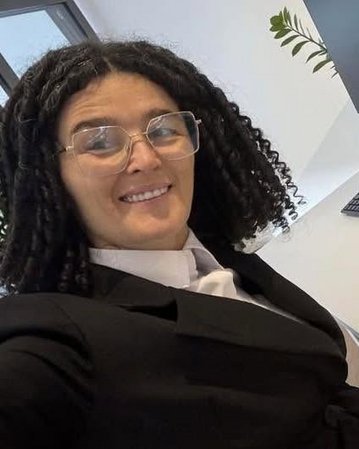 Eine Person mit lockigem Haar und Brille trägt einen Anzug und sitzt an einem Schreibtisch mit einem Computer und einer Tastatur.