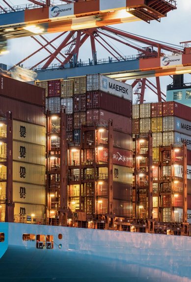 Containerschiff mit zahlreichen beladenen Containern im Hafen, umgeben von Kränen und Hafeninfrastruktur.