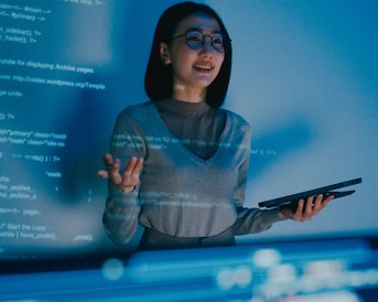 Eine Frau mit Brille steht vor einem Bildschirm, auf dem Programmiercode angezeigt wird. Sie hält ein Tablet in der Hand und erklärt etwas, während sie lächelt.