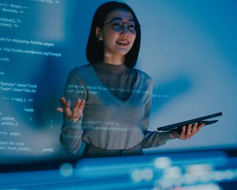 Eine Frau mit Brille steht vor einem Bildschirm, auf dem Programmiercode angezeigt wird. Sie hält ein Tablet in der Hand und erklärt etwas, während sie lächelt.