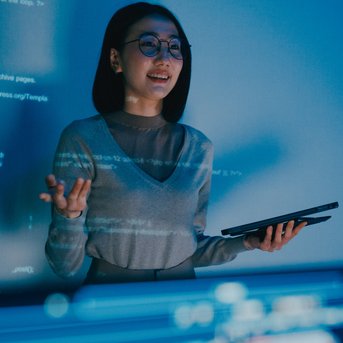 A woman with glasses stands in front of a screen displaying programming code. She is holding a tablet in her hand and explaining something while smiling.