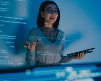 A woman with glasses stands in front of a screen displaying programming code. She is holding a tablet in her hand and explaining something while smiling.