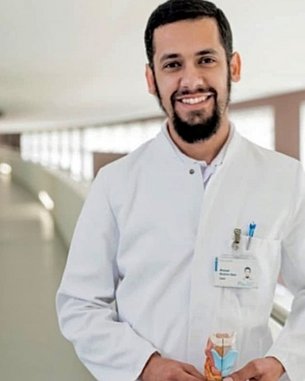 Ein Arzt in einem weißen Kittel hält ein anatomisches Modell in der Hand und lächelt. Der Hintergrund zeigt einen modernen Krankenhausflur.