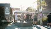 Menschen stehen vor einem Food Truck und warten auf Essen. Einige unterhalten sich, während andere in einer Schlange stehen.