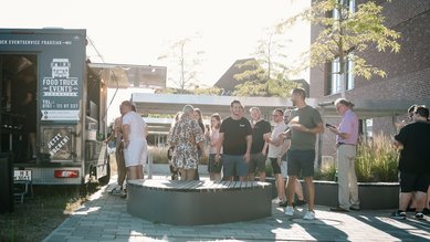 Menschen stehen vor einem Food Truck und warten auf Essen. Einige unterhalten sich, während andere in einer Schlange stehen.