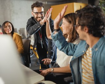 A group of people in a modern office is giving each other high-fives while working at their computers.