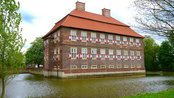 Historisches Gebäude mit rotem Dach und bunten Fahnen, umgeben von Wasser und Bäumen.