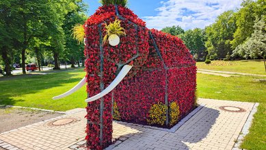 Ein mit Blumen gestalteter Elefant steht in einem Park, umgeben von Bäumen und Rasenflächen. Die Struktur hat Stoßzähne aus Kunststoff.