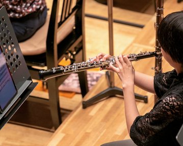 Eine Musikerin spielt Oboe, während sie auf ein digitales Notenblatt schaut. Im Hintergrund sind weitere Musiker und Stühle zu sehen.