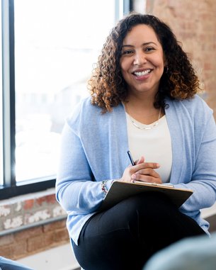 Eine Frau mit lockigem Haar sitzt lächelnd in einer Besprechung, hält einen Notizblock und spricht mit zwei Personen.