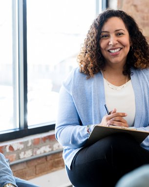 Eine Frau mit lockigem Haar sitzt lächelnd in einer Besprechung, hält einen Notizblock und spricht mit zwei Personen.
