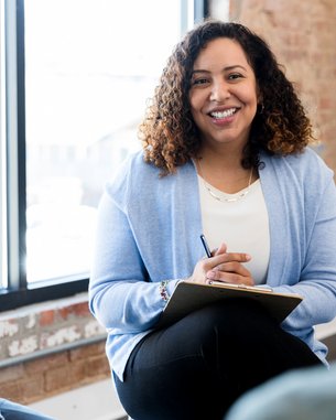 Eine Frau mit lockigem Haar sitzt lächelnd in einer Besprechung, hält einen Notizblock und spricht mit zwei Personen.