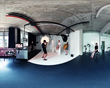 Ein Studio mit einem runden Fotohintergrund, einem Fotografen, der ein Model ablichtet. Im Hintergrund sind weitere Personen und Ausrüstungen sichtbar. Die Wände sind hell, und der Boden ist dunkel.