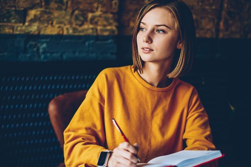Eine junge Frau in einem orangefarbenen Pullover sitzt an einem Tisch, hält einen Stift und schreibt in ein Notizbuch.