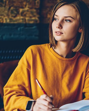 Eine junge Frau in einem orangefarbenen Pullover sitzt an einem Tisch, hält einen Stift und schreibt in ein Notizbuch.