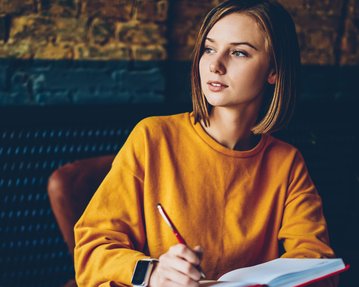 Eine junge Frau in einem orangefarbenen Pullover sitzt an einem Tisch, hält einen Stift und schreibt in ein Notizbuch.