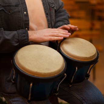 Ein Mann spielt auf zwei Congas, während er auf einem Stuhl sitzt. Er trägt ein offenes Hemd und hat die Hände auf den Trommeln. Im Hintergrund ist ein Notenständer mit Musiknoten sichtbar.