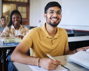 A young man with glasses sits at a desk in a classroom, smiling and holding a book. In the background, two other students can be seen sitting at their desks as well.