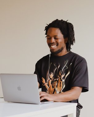 A man with curly hair is sitting at a table, working on a laptop. He is wearing a T-shirt with an artistic design. Next to him, there is a cup. The room has a light, neutral wall color.