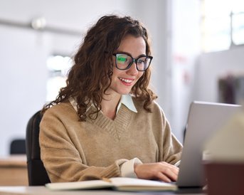 Eine lächelnde Frau mit lockigem Haar und Brille sitzt an einem Schreibtisch und arbeitet an einem Laptop. Sie trägt einen beigen Pullover und scheint konzentriert zu sein.