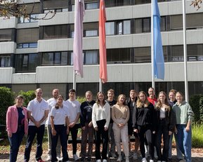 A group of 18 people stands in front of a building, surrounded by flags. The participants are wearing various casual outfits and smiling.