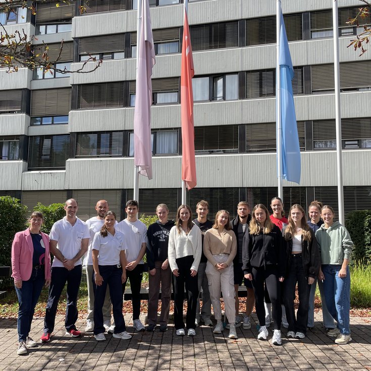 A group of 18 people stands in front of a building, surrounded by flags. The participants are wearing various casual outfits and smiling.