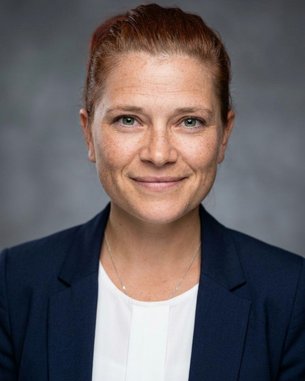 Porträt einer Frau mit roten Haaren, die einen blauen Blazer trägt. Sie hat grüne Augen und lächelt leicht.