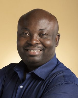 A man with a bald head and a broad smile is wearing a blue shirt. The background is in soft shades of gold.