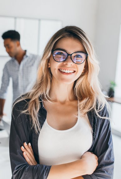 A smiling woman with glasses stands in the foreground, while in the background, several people are working at a table and talking to each other. The room is bright and modern in design.