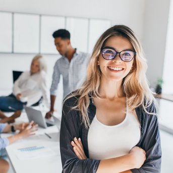 Eine lächelnde Frau mit Brille steht im Vordergrund, während im Hintergrund mehrere Personen an einem Tisch arbeiten und miteinander sprechen. Der Raum ist hell und modern gestaltet.