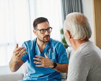 A doctor in a blue uniform is talking to an older man. The doctor is gesturing as he provides information or advice. Both individuals are sitting in a bright room.