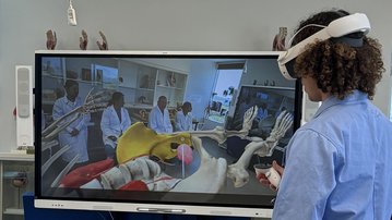 Ein Student mit einer VR-Brille steht vor einem Bildschirm, der eine medizinische Demonstration mit mehreren Personen zeigt.
