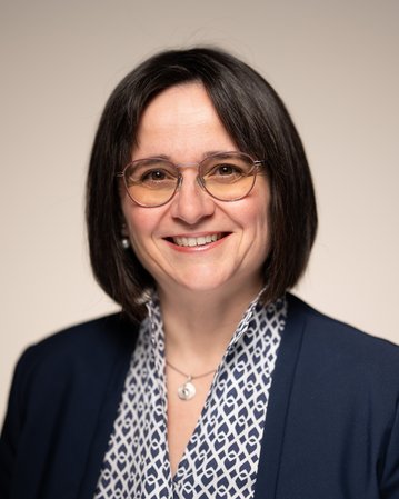 Eine Frau mit schulterlangen, dunklen Haaren und Brille, trägt einen blauen Blazer und ein gemustertes Oberteil, lächelt freundlich.