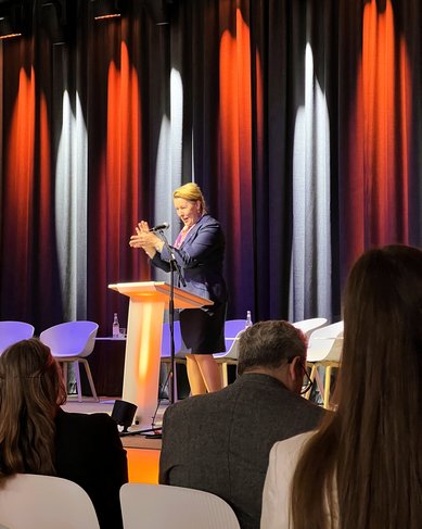 Eine Frau steht am Rednerpult und spricht, während sie ihre Hände gestikuliert. Im Hintergrund sind Stühle und eine Bühne mit orangefarbenen und schwarzen Vorhängen sichtbar. Einige Zuhörer sind im Vordergrund zu sehen.