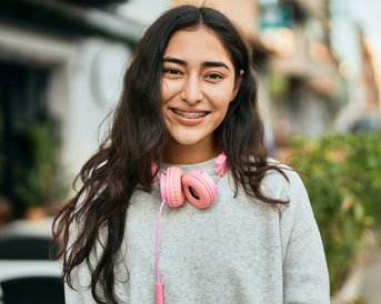 Ein lächelndes Mädchen mit Brackets trägt rosa Kopfhörer und ein graues Sweatshirt. Im Hintergrund sind verschwommene Straßenszenen und Pflanzen zu sehen.