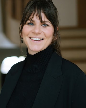 A smiling woman with long, dark hair is wearing a black turtleneck sweater and a blazer. She is standing in a bright interior.