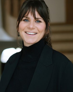Eine lächelnde Frau mit langen, dunklen Haaren trägt einen schwarzen Rollkragenpullover und einen Blazer. Sie steht in einem hellen Innenraum.