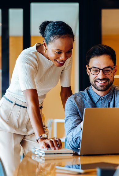Two people in a modern office are looking at a laptop. A woman is standing behind a man, smiling as he works.