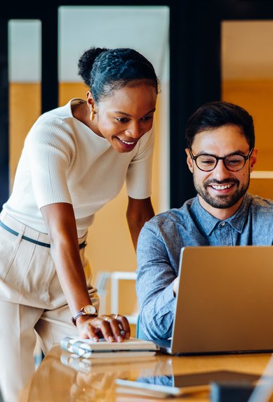 Zwei Personen in einem modernen Büro schauen auf einen Laptop. Eine Frau steht hinter einem Mann und lächelt, während er arbeitet.