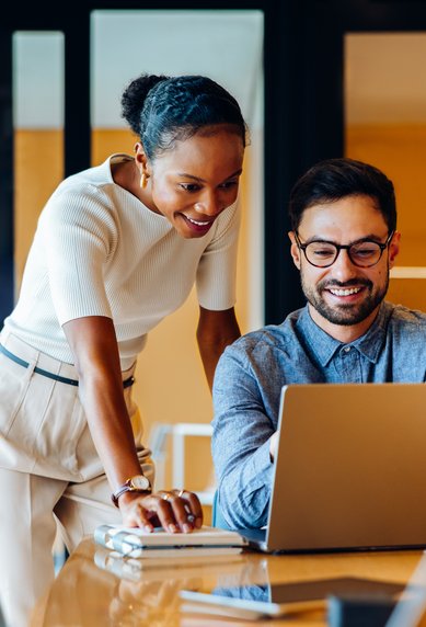 Zwei Personen in einem modernen Büro schauen auf einen Laptop. Eine Frau steht hinter einem Mann und lächelt, während er arbeitet.