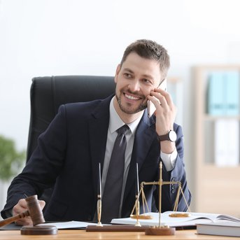 Ein Anwalt sitzt an einem Schreibtisch, telefoniert und lächelt. Auf dem Tisch befinden sich eine Richterglocke und eine Waage.