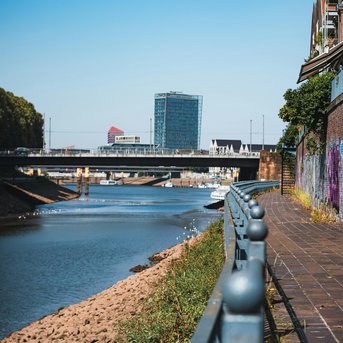 Uferpromenade entlang eines Flusses mit einer Brücke im Hintergrund und modernen Gebäuden in der Ferne.