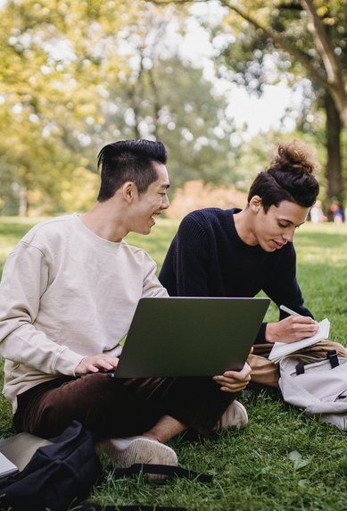 Two people are sitting on the grass in the park. One person is working on a laptop while the other is taking notes. Surrounding them are trees and a relaxed atmosphere.