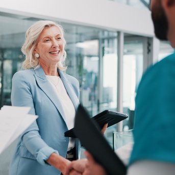 Eine lächelnde Frau in einem blauen Blazer schüttelt die Hand eines Mannes, während eine andere Person mit Unterlagen daneben steht.