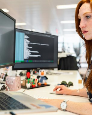 A woman with long red hair is sitting at a desk, working on a laptop. In front of her are two large monitors displaying programming code. The workspace is modern and well-organized.